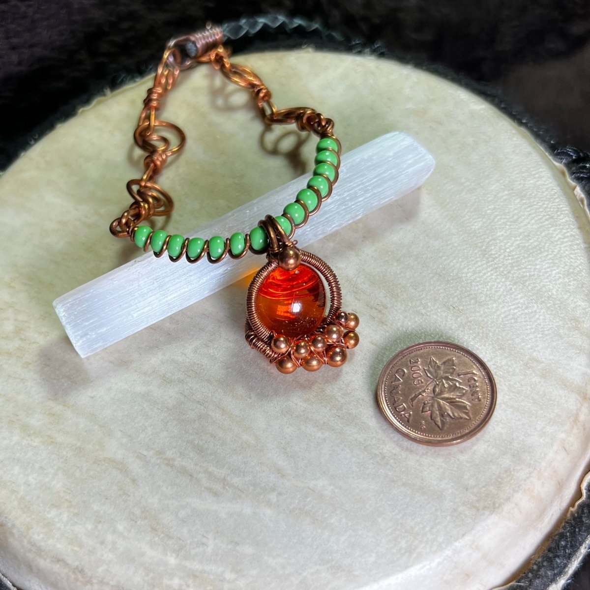 Necklace with green beads and an orange glass cabochon wrapped in raw copper, on a white surface, next to a penny for scale.