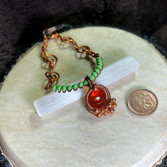 Necklace with green beads and an orange  glass cabochon wrapped in raw copper, on a white surface, next to a penny for scale.