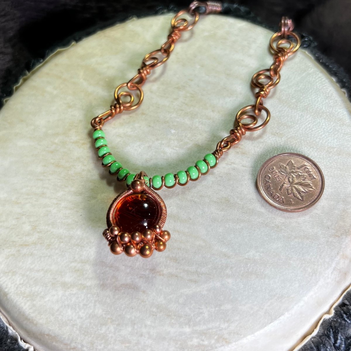Necklace with green beads and an orange glass cabochon wrapped in raw copper, on a white surface, next to a penny for scale.