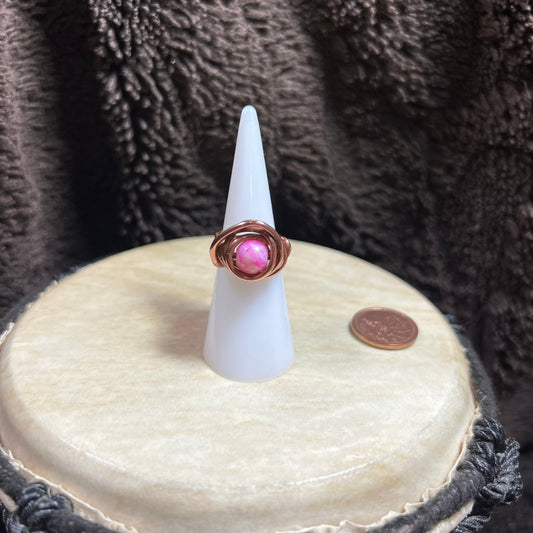 Ring with pink glass bead on a white cone stand against a textured brown background, next to a penny for scale.