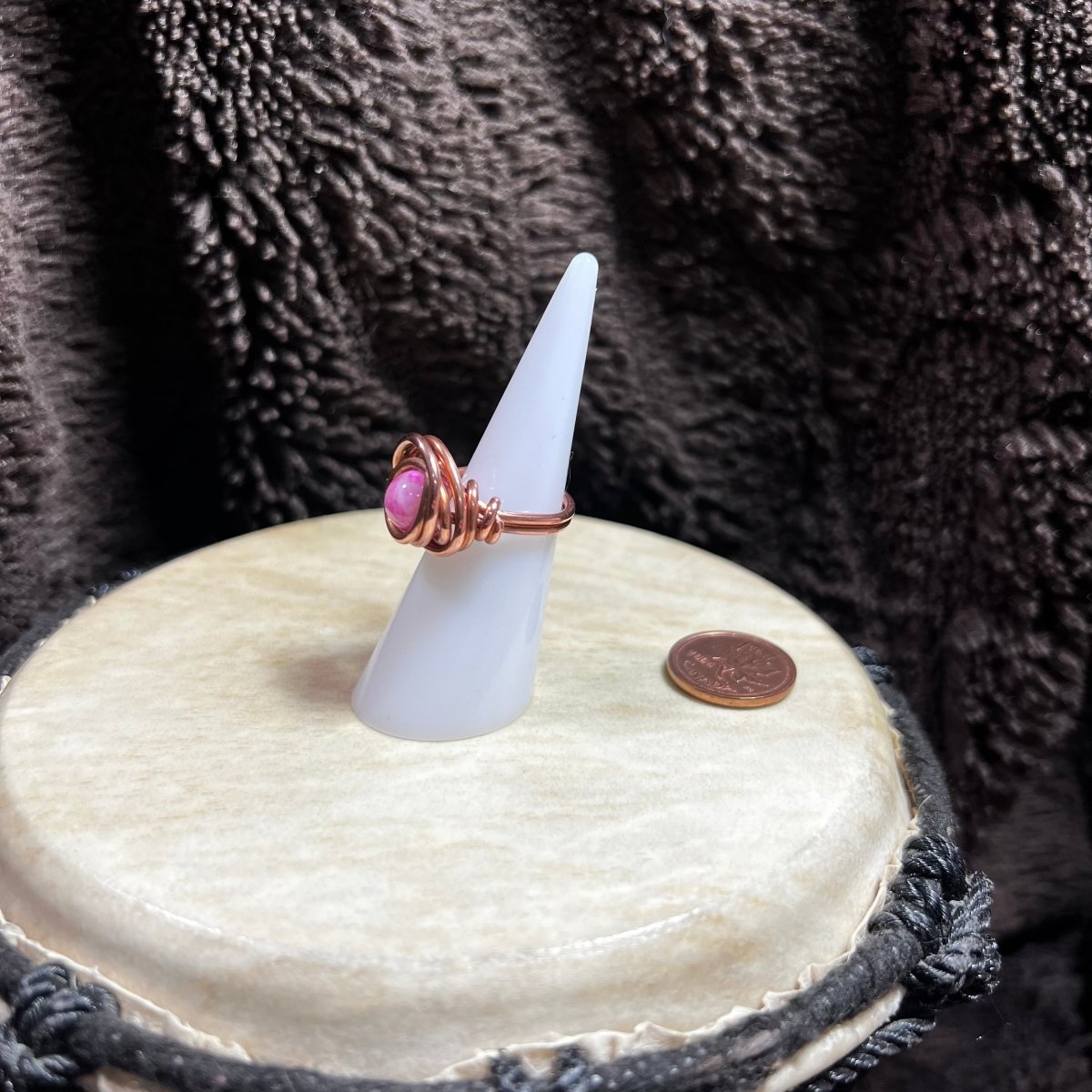 Ring with pink glass bead on a white cone stand against a textured brown background, next to a penny for scale.