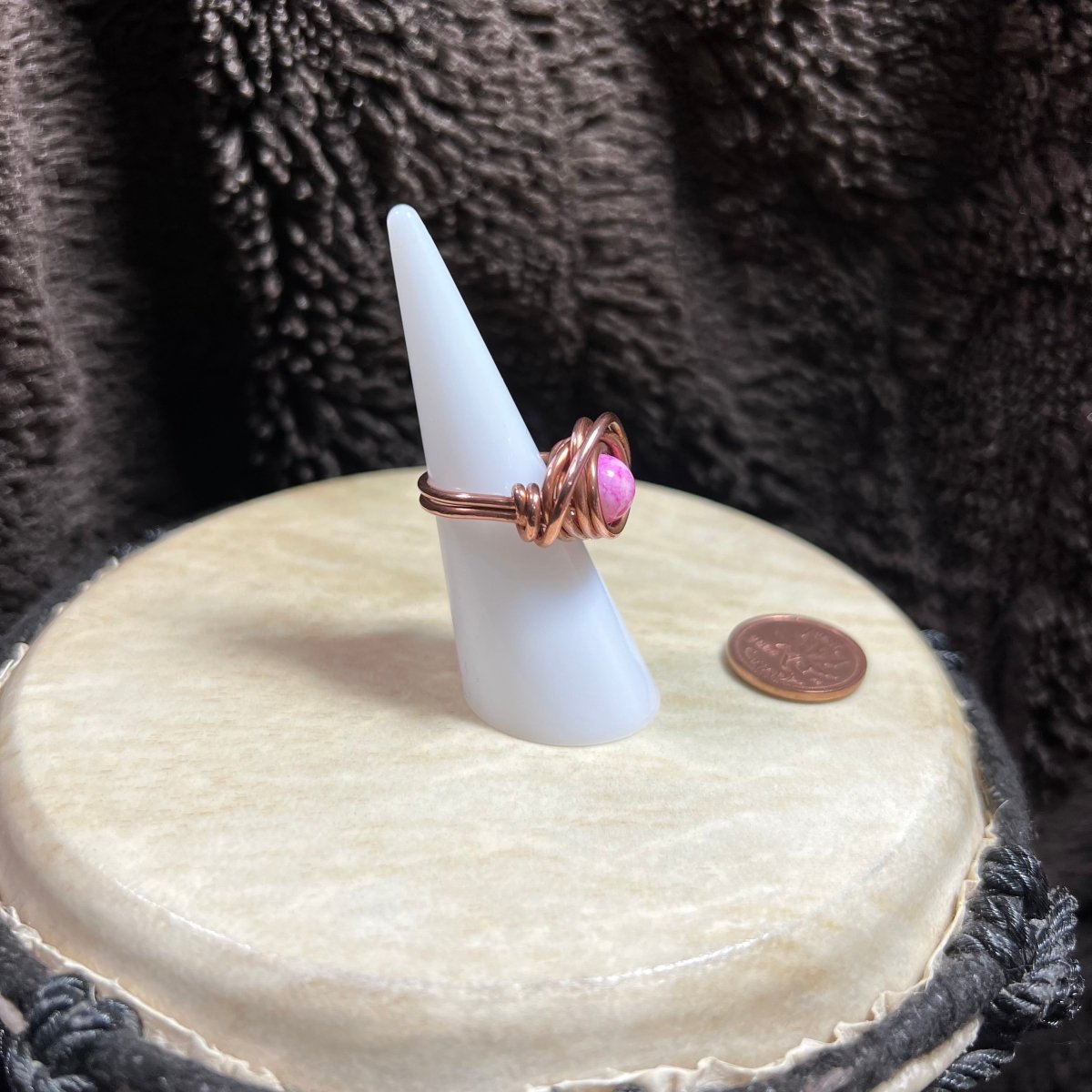 Ring with pink glass bead on a white cone stand against a textured brown background, next to a penny for scale.