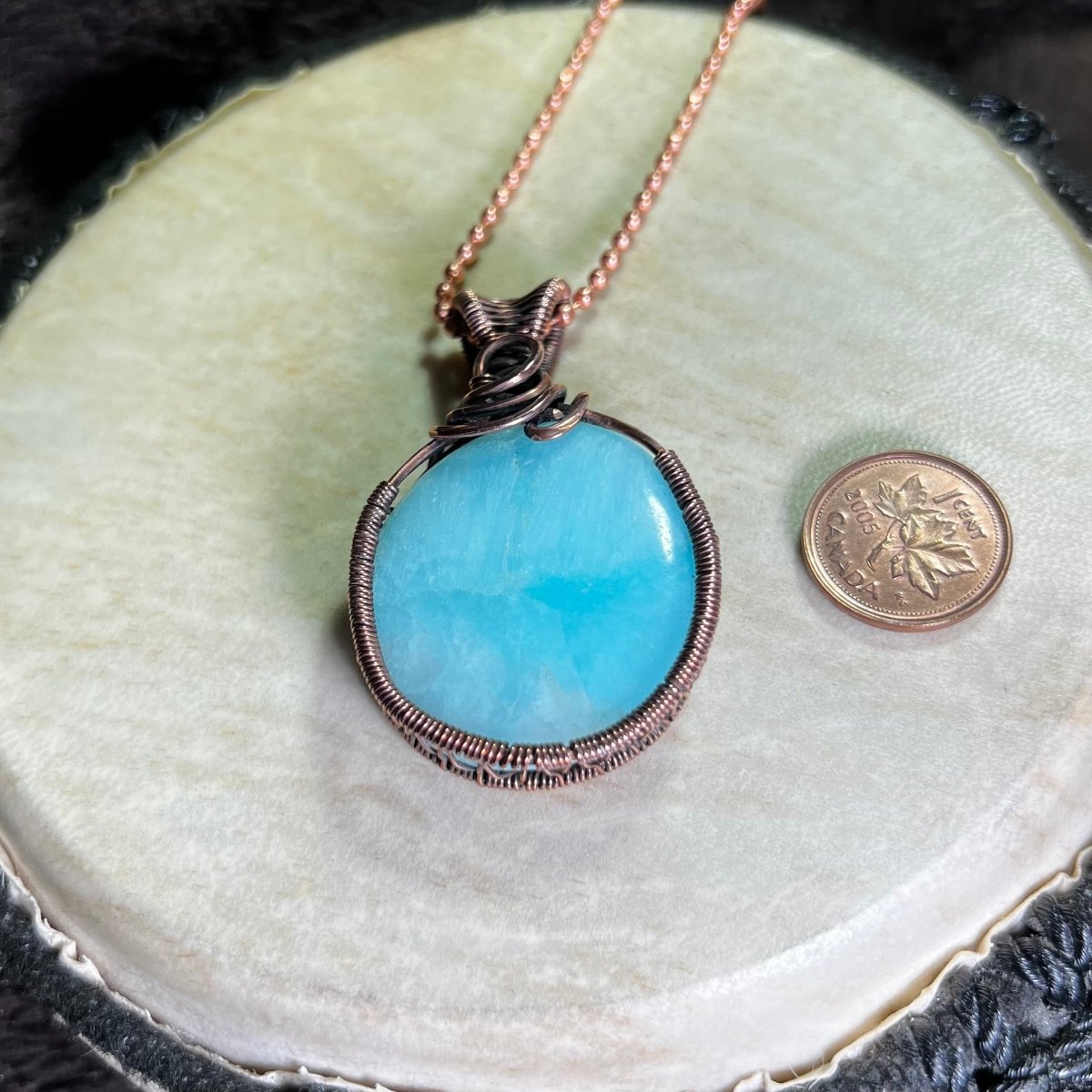 Blue aragonite pendant with copper wire wrapping on a white surface next to a penny for scale