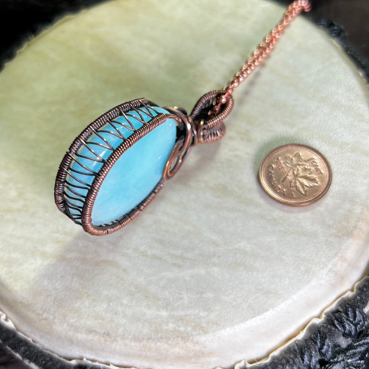 Blue aragonite pendant with copper wire wrapping on a white surface next to a penny for scale