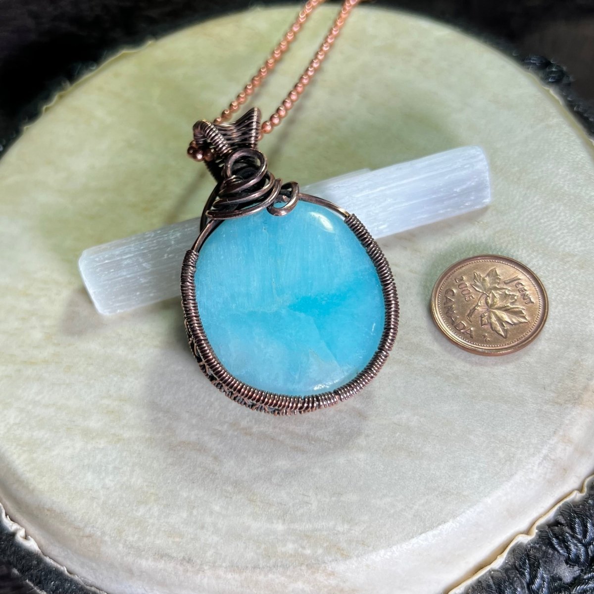 Blue aragonite pendant with copper wire wrapping on a white surface next to a penny for scale