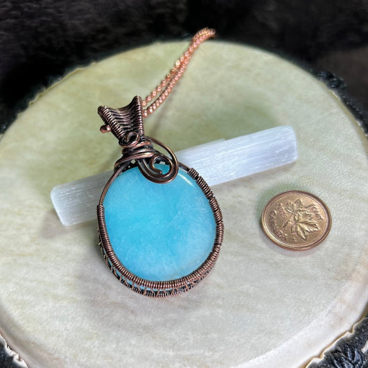 Blue aragonite pendant with copper wire wrapping on a white surface next to a penny for scale