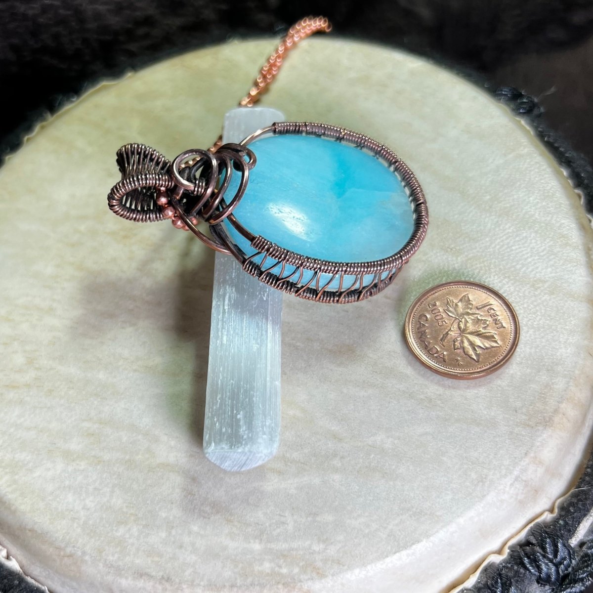 Blue aragonite pendant with copper wire wrapping on a white surface next to a penny for scale