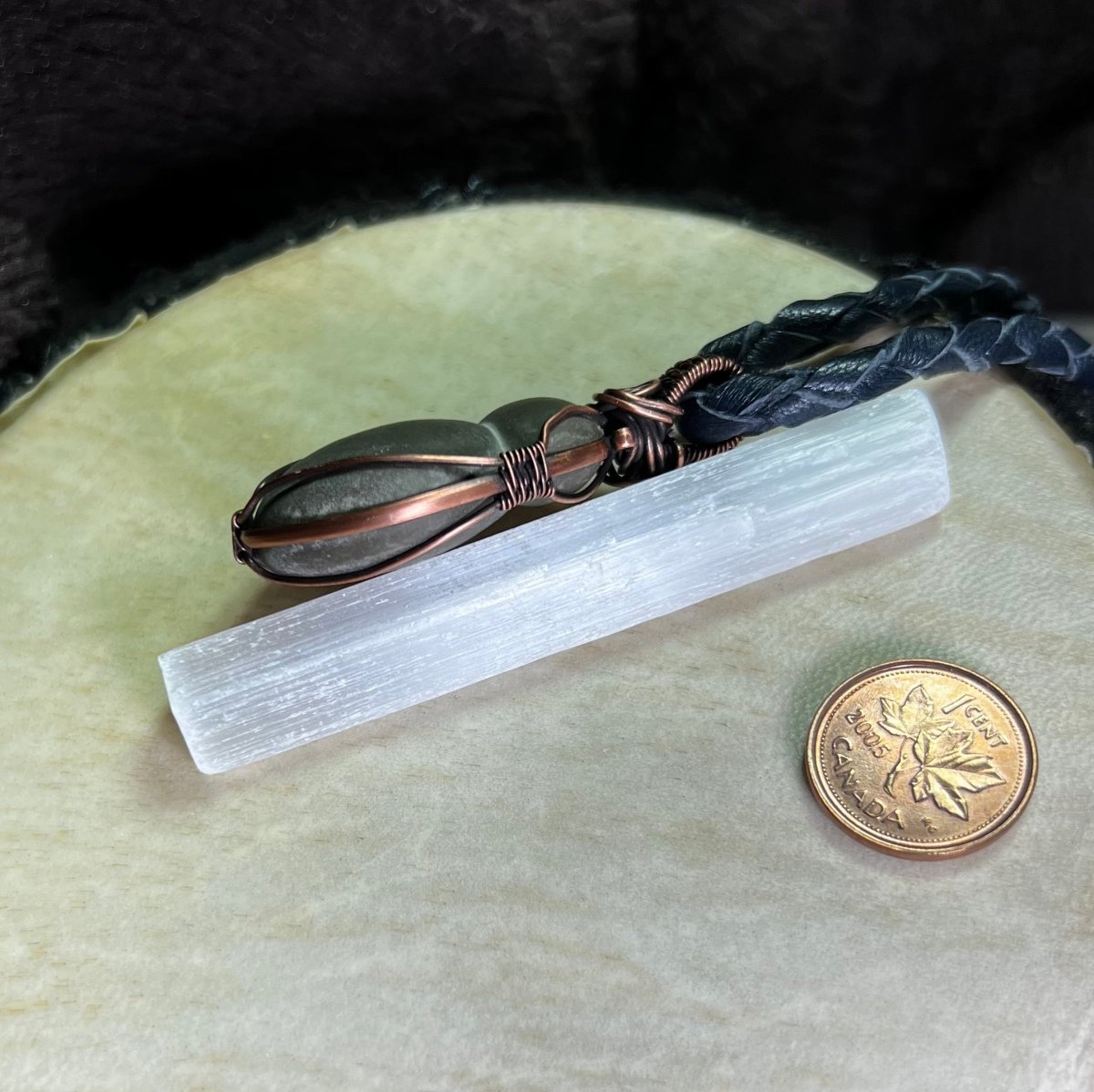 Side views of a necklace with a bulbous teardrop-shaped Canadian Fairy Stone wrapped in copper on a cream surface, next to a penny for scale.