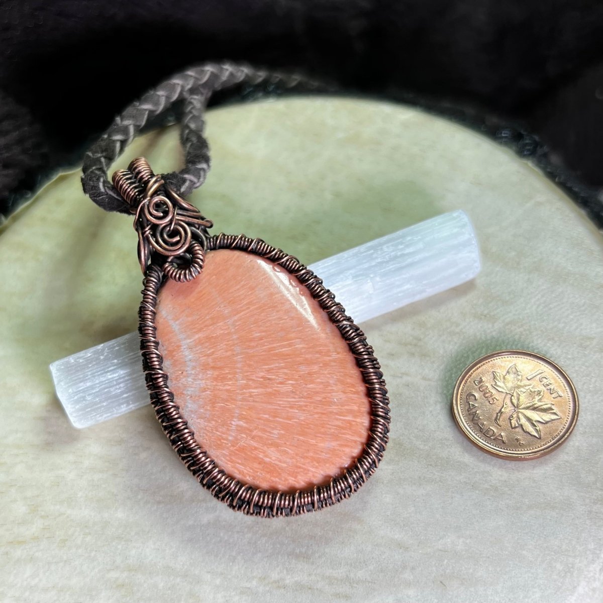 Necklace with a peach celestobarite stone wrapped in copper on a cream surface next to a penny for scale.