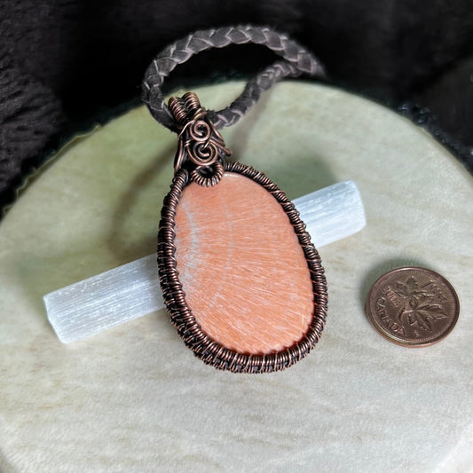 Necklace with a peach celestobarite stone wrapped in copper on a cream surface next to a penny for scale.