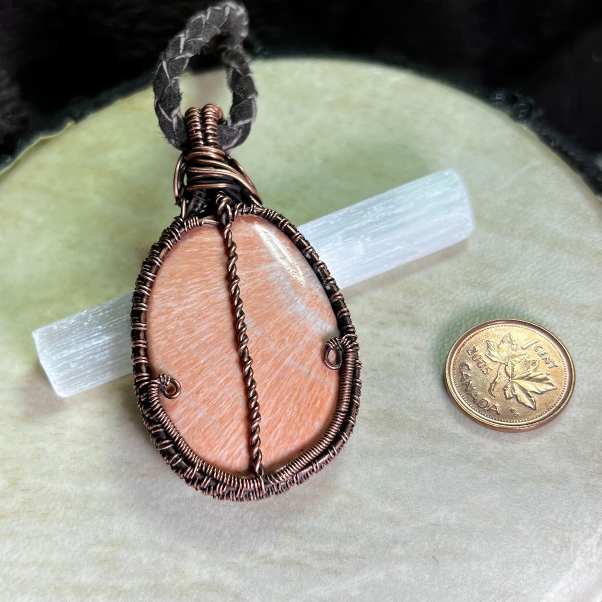 Back view of a necklace with a peach celestobarite stone wrapped in copper on a cream surface next to a penny for scale.
