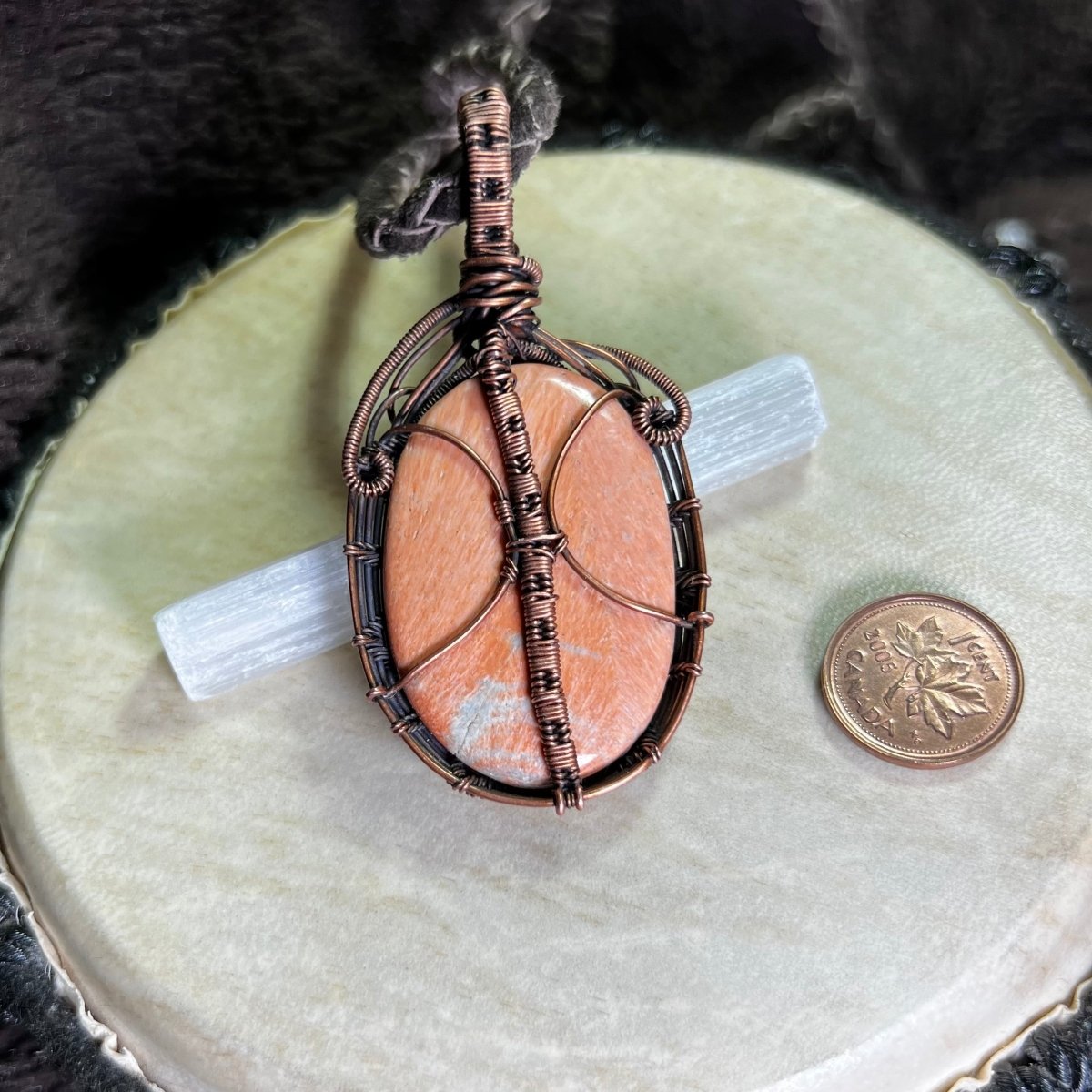 Back side of a copper-wrapped stone pendant on a beige surface with a penny for scale.