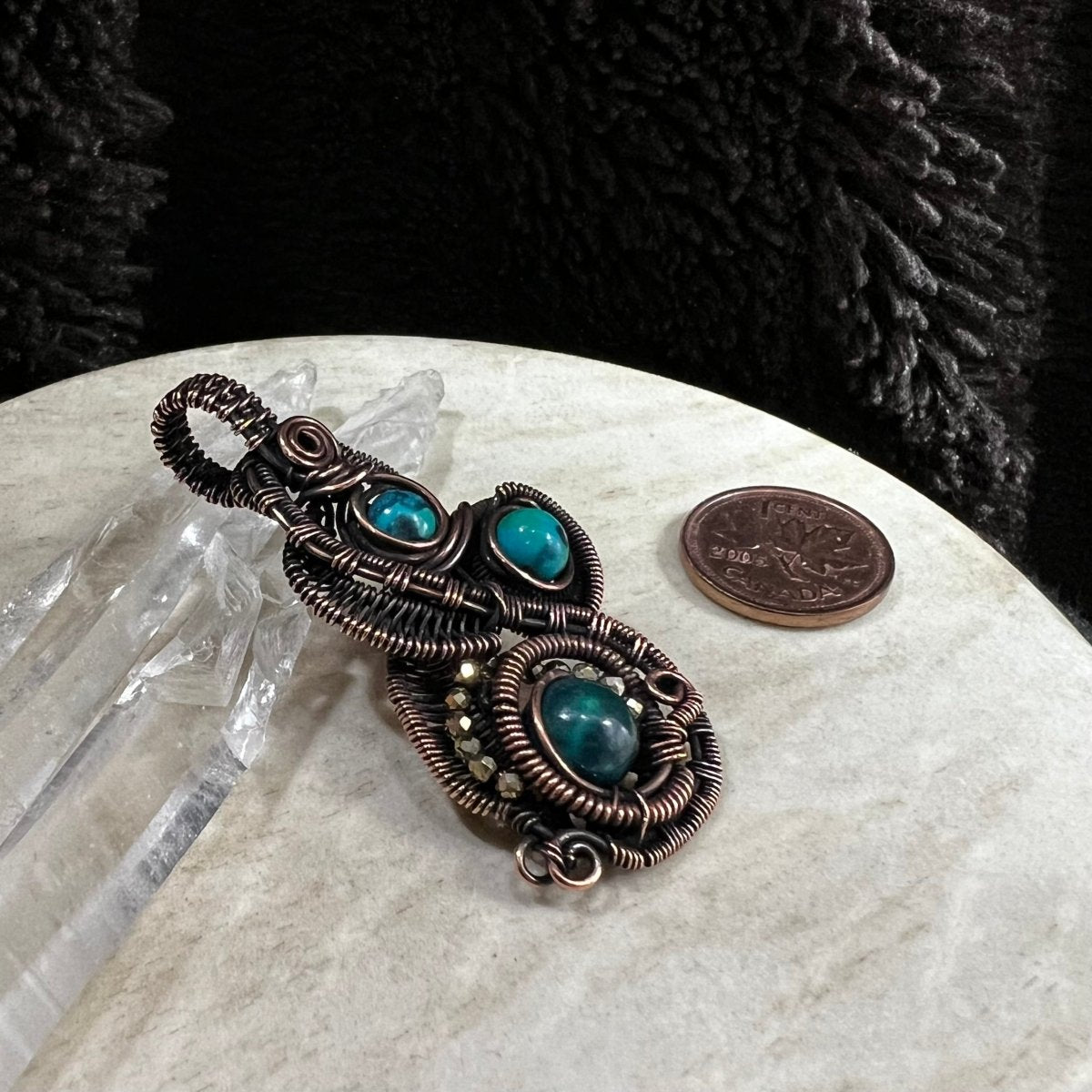 Antiqued copper intricate wirework pendant with chrysocolla beads on a white surface next to a Canadian penny for scale.
