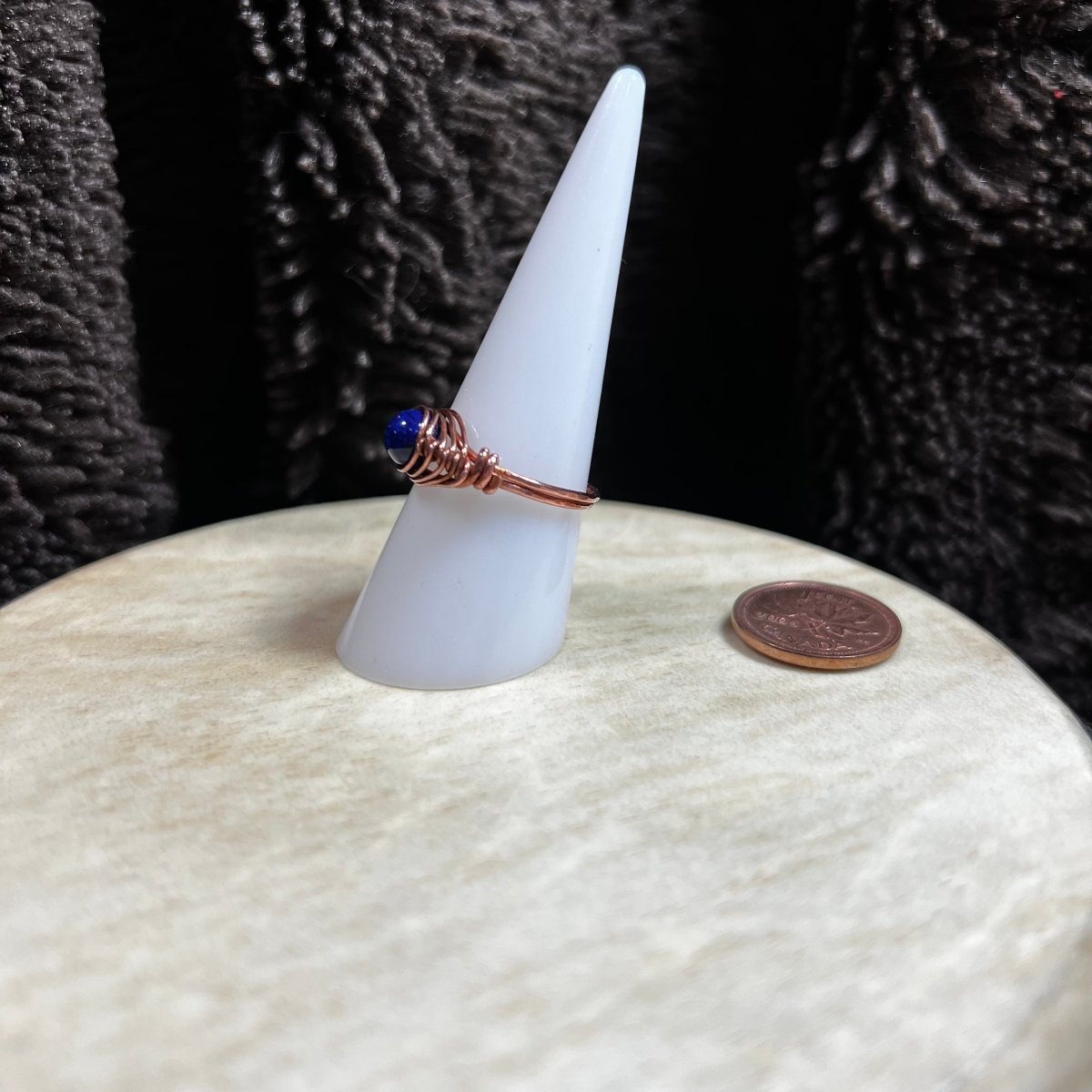 Ring with a blue stone on a white cone-shaped stand, next to a penny for scale, against a dark textured background.