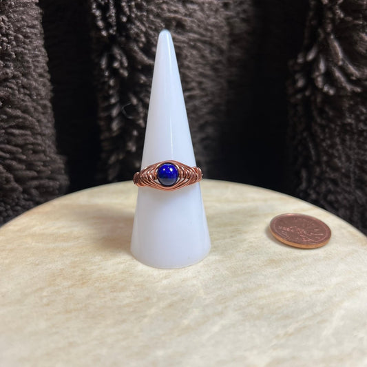 Ring with a blue stone on a white cone-shaped stand, next to a penny for scale, against a dark textured background.