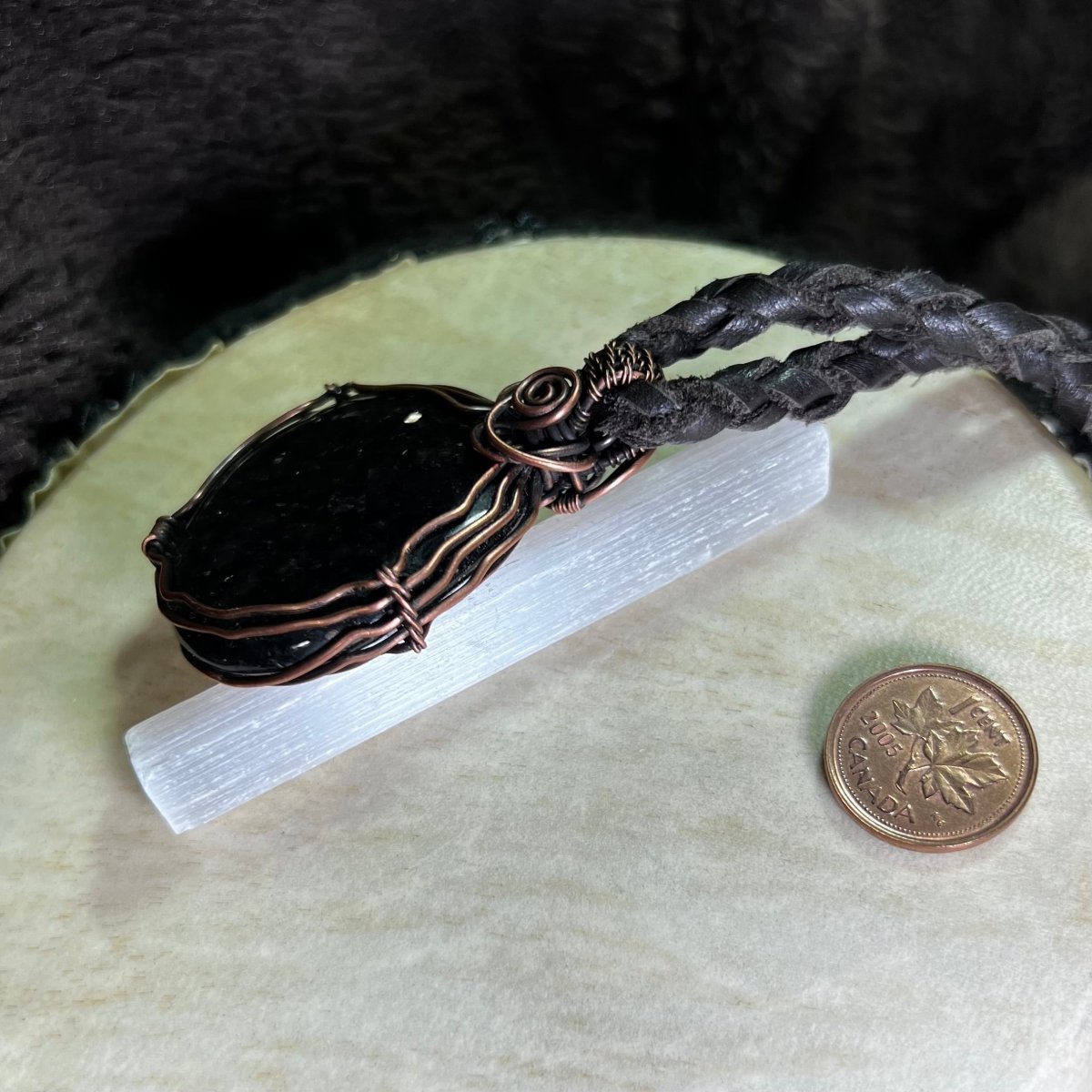 Black coppernite stone pendant in a copper wire setting on a white surface next to a penny for scale.
