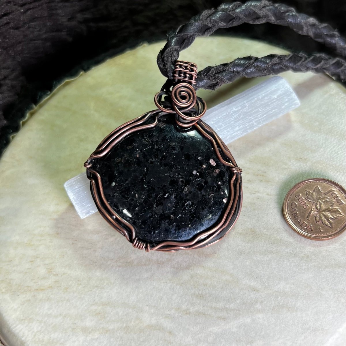 Black coppernite stone pendant in a copper wire setting on a white surface next to a penny for scale.