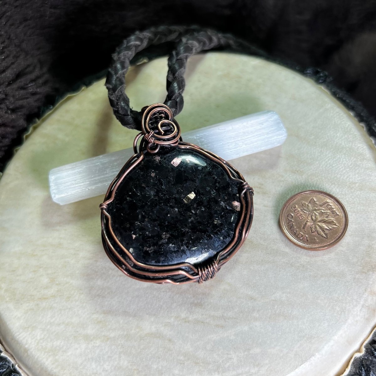 Black coppernite stone pendant in a copper wire setting on a white surface next to a penny for scale.