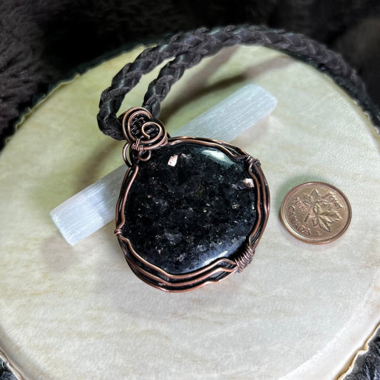 Black coppernite stone pendant in a copper wire setting on a white  surface next to a penny for scale.