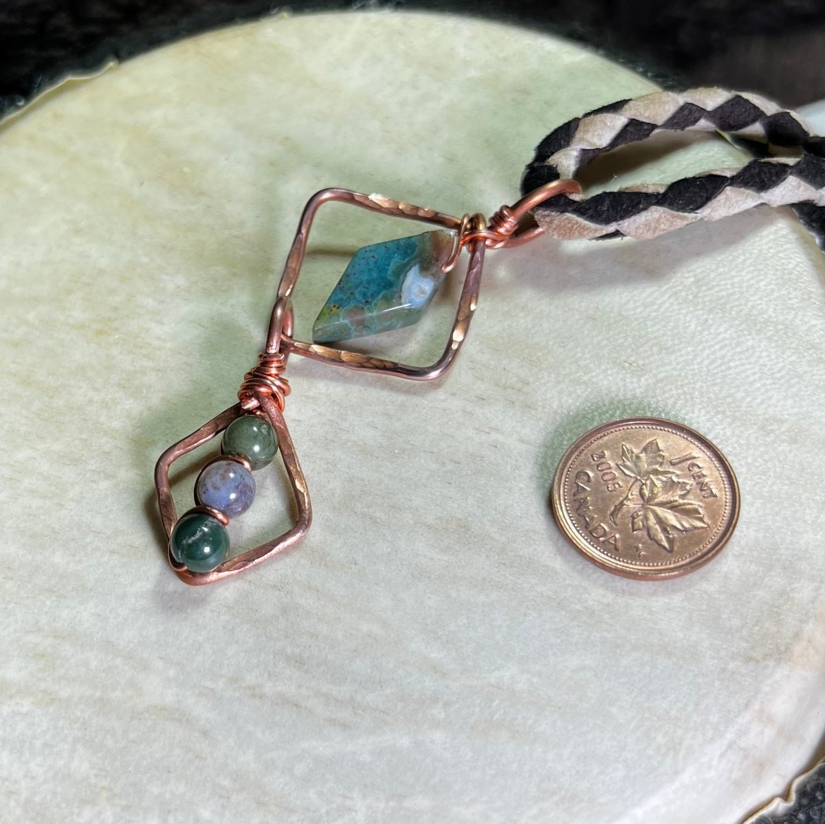 Necklace with geometric copper design and moss agate stones on a white surface, next to a penny for scale