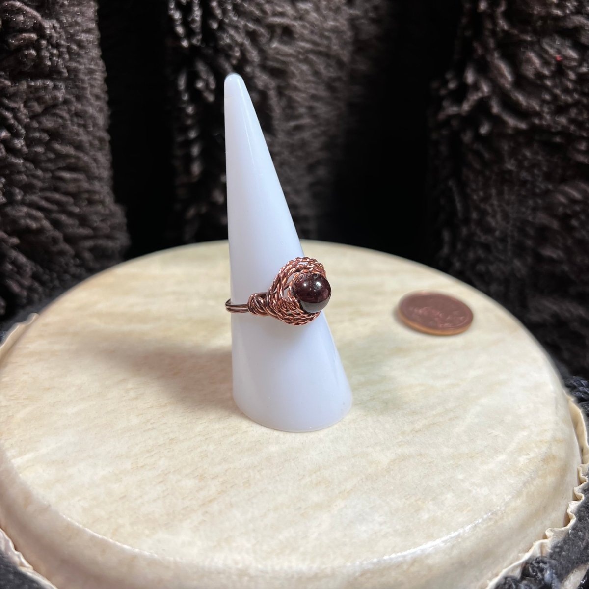 Ring with deep red garnet bead on a white cone-shaped stand against a textured dark background