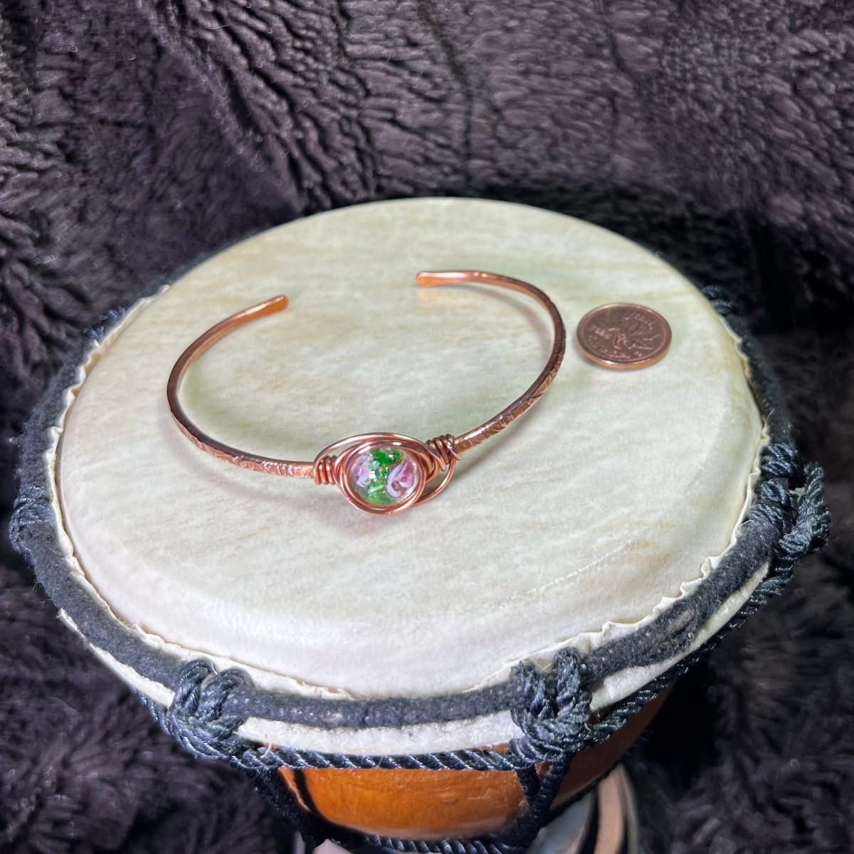 Copper bracelet with a lampwork focal on a round surface next to a penny for scale and a dark textured background