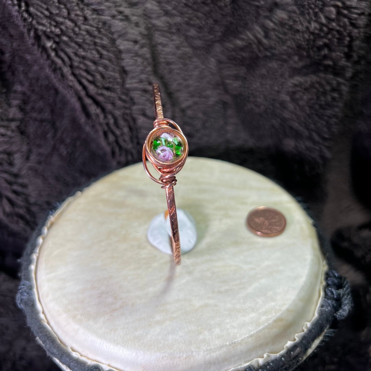Copper bracelet with a lampwork focal on a round white surface next to a penny for scale against a dark textured background