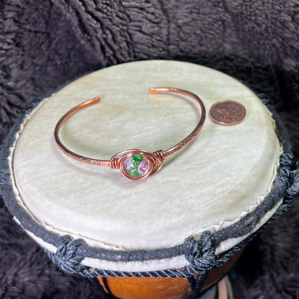 Copper bracelet with a lampwork bead on a round surface next to a penny for scale with a dark textured background