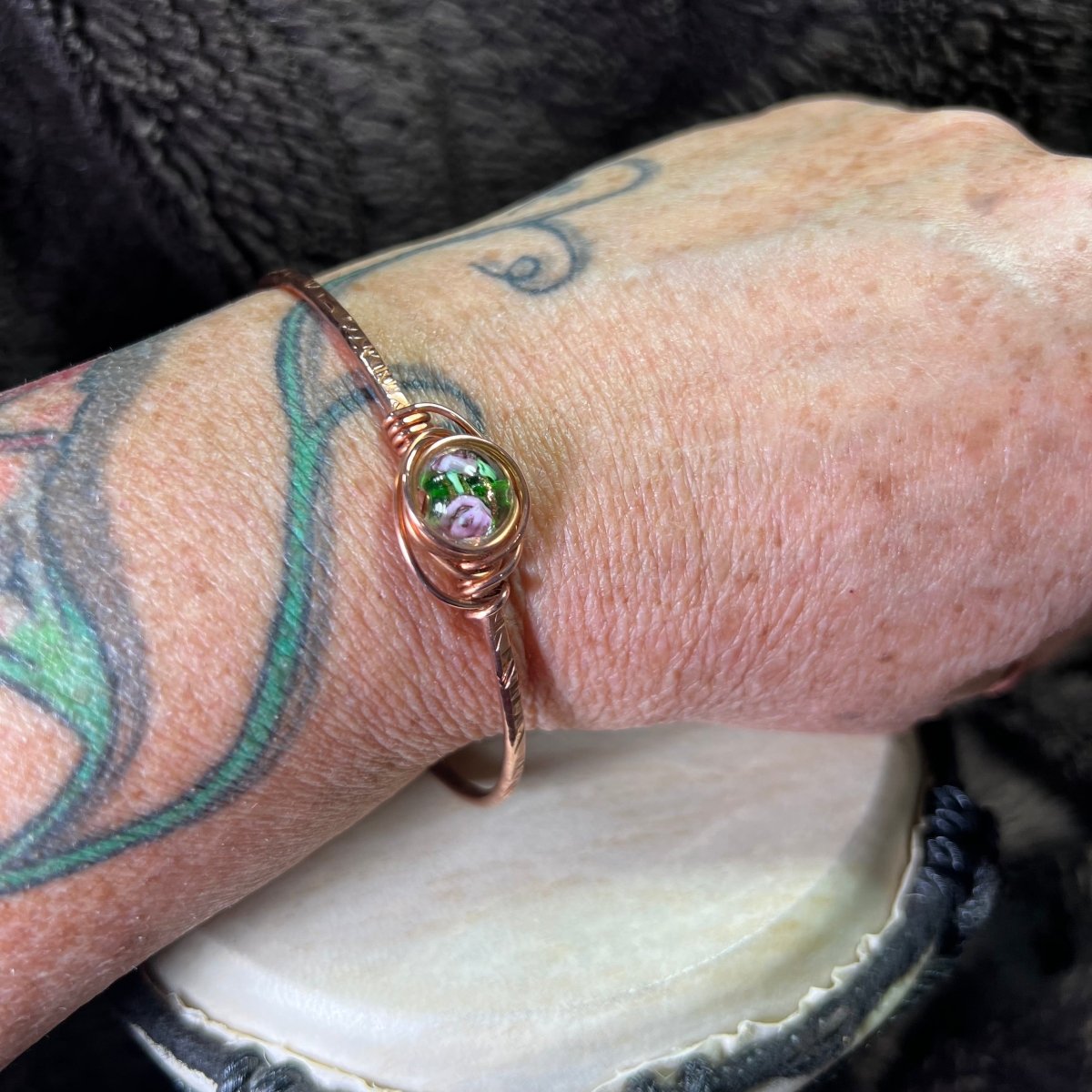 Copper bracelet with a green and pink lampwork focal on a wrist with tattoos.