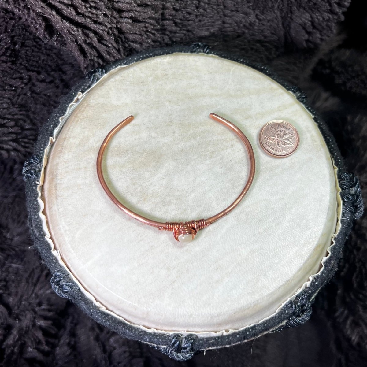 Copper bracelet on a round white surface next to a penny for scale and a dark textured background