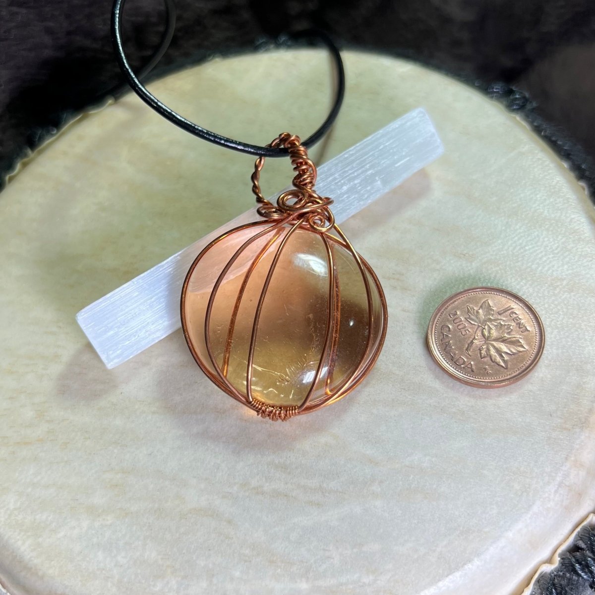 Copper wire-wrapped glass pumpkin on a black cord next to a penny for scale on a white surface