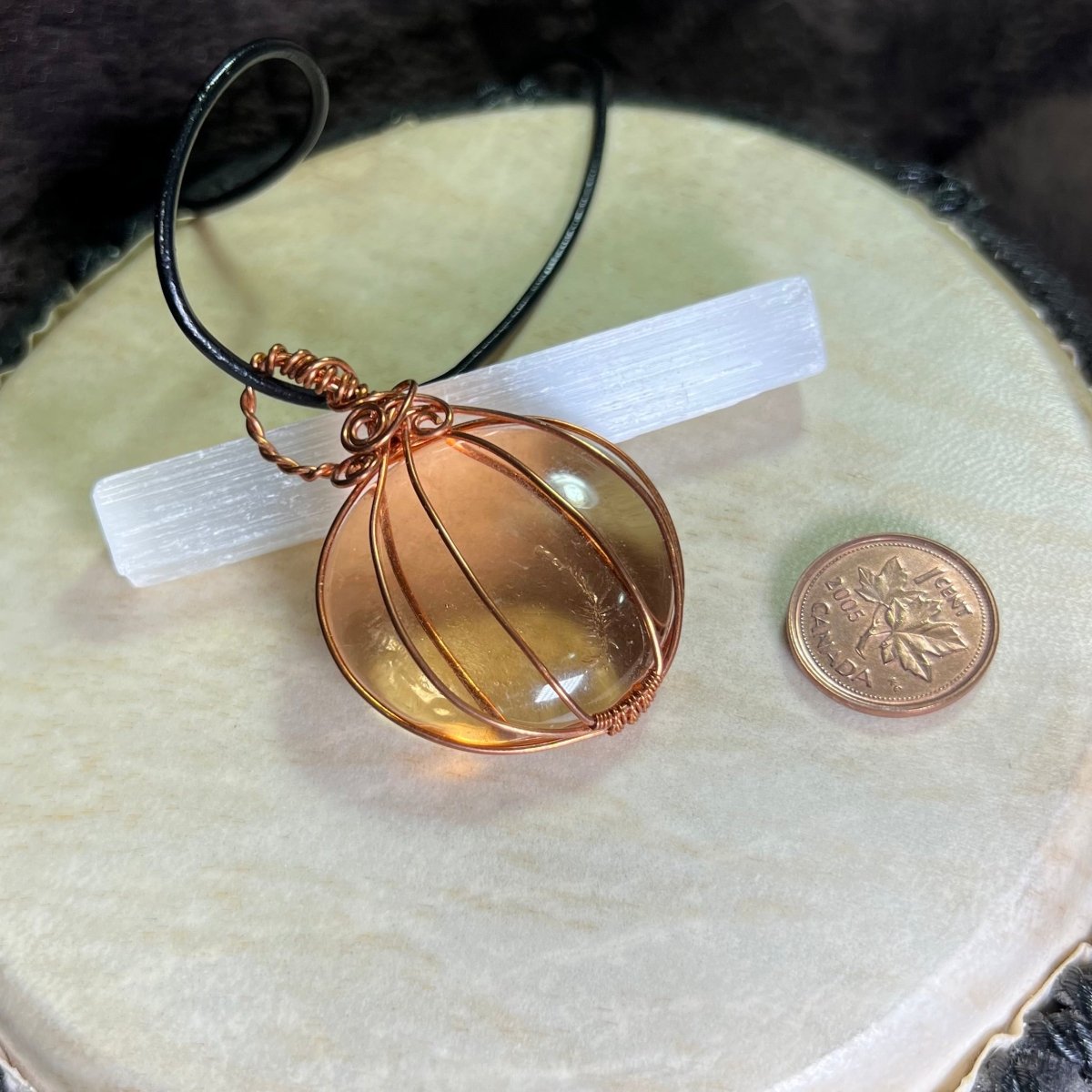 Copper wire-wrapped glass pumpkin on a black cord next to a penny for scale on a white surface