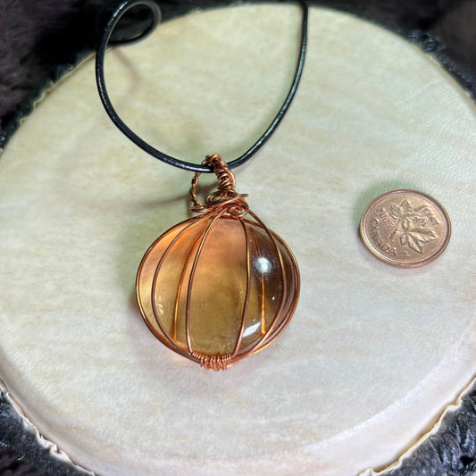 Copper wire-wrapped glass pumpkin on a black cord next to a penny for scale on a white surface