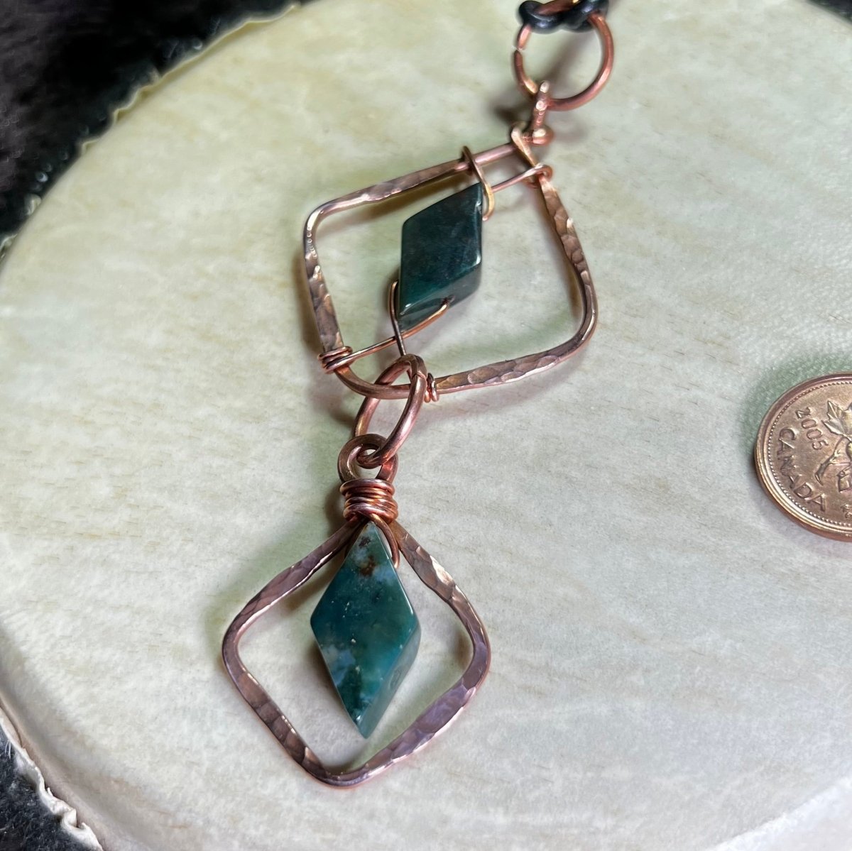 Copper 2 piece diamond-shaped pendant with moss agate gemstones on a beige surface with a penny for scale.