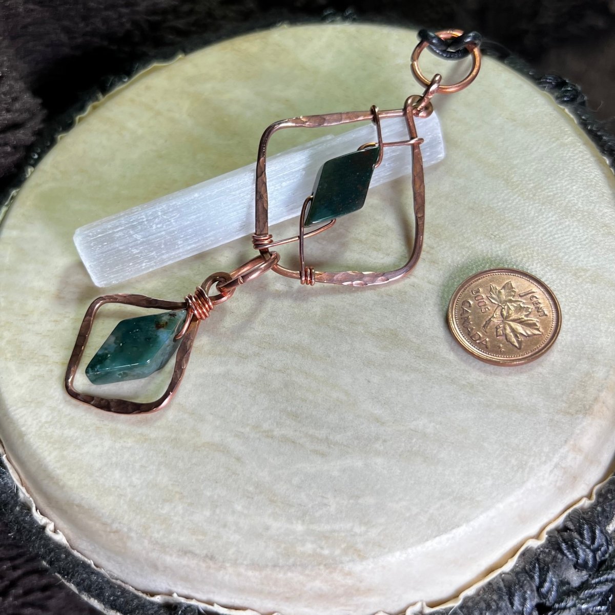 Copper 2 piece diamond-shaped pendant with moss agate gemstones leaning on a selenite stick on a beige surface with a penny for scale.