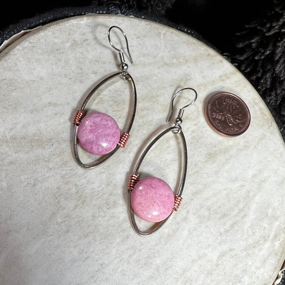 Handmade rhodochrosite bead earrings suspended in reclaimed silver-plated oval hoops with copper wire accents and surgical steel ear wires, photographed with Canadian penny for scale.