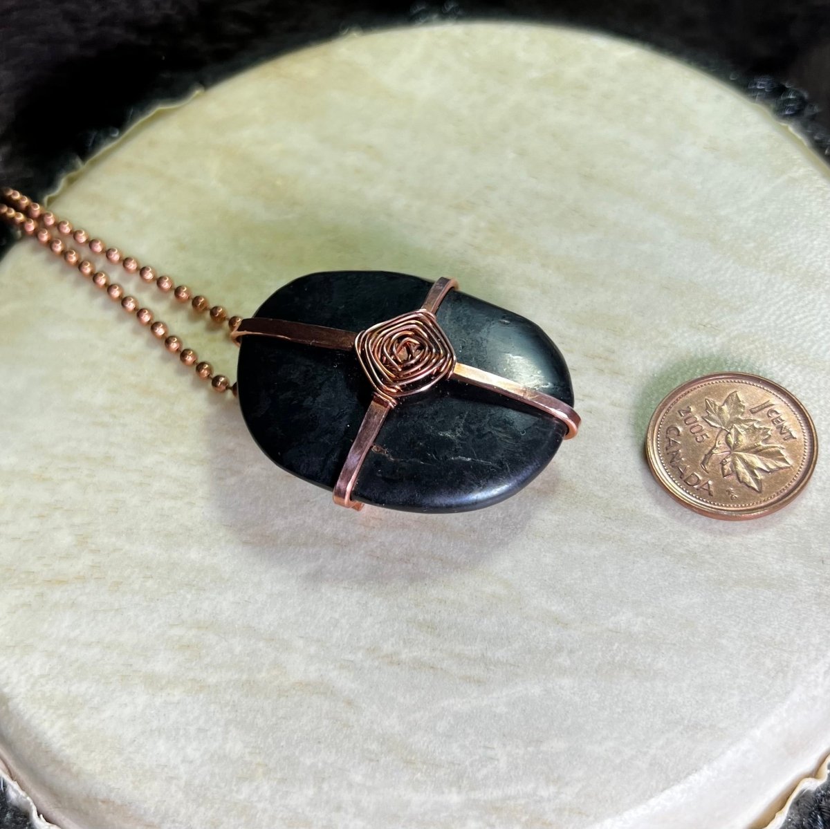 Raw Copper necklace with a black Shungite stone in a God's Eye wrap on a white surface next to a penny for scale.