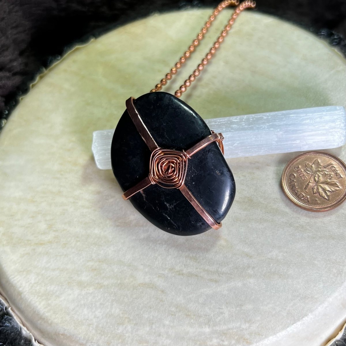 Raw Copper necklace with a black Shungite stone in a God's Eye wrap on a white surface next to a penny for scale.