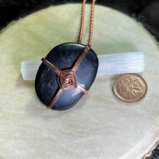 Raw Copper necklace with a black Shungite stone in a God's Eye wrap on a white surface next to a penny for scale.