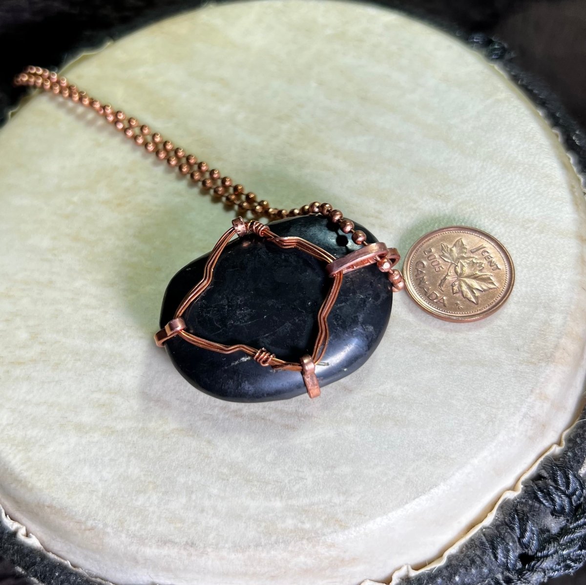 Back side view of a raw Copper necklace with a black Shungite stone on a white surface next to a penny for scale.