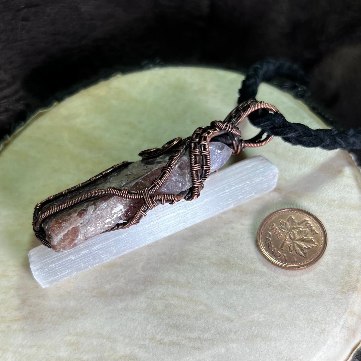 Side view of a Super Seven Crystal pendant with copper wire wrapping on a cream surface next to a penny for scale.