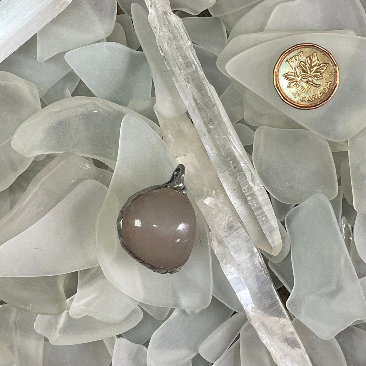 Bay of Fundy Quartz Pendant top down view. In a bed of sea glass and quartz crystals, next to a Canadian penny for scale.
