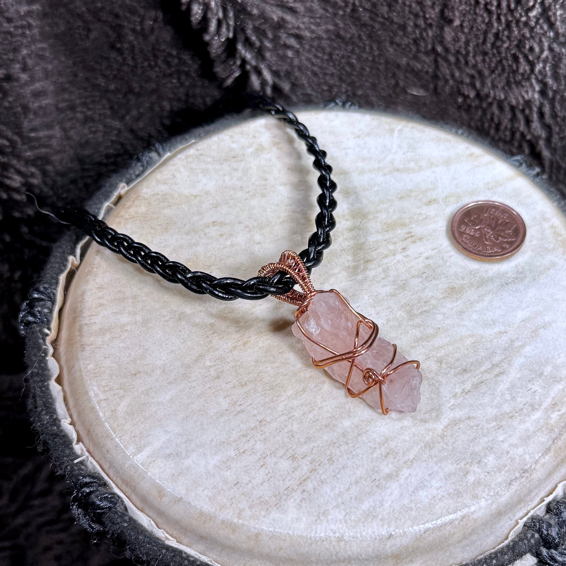Raw Rose Quartz crystal wrapped in reclaimed copper on black braided leather, displayed with a Canadian penny for scale.