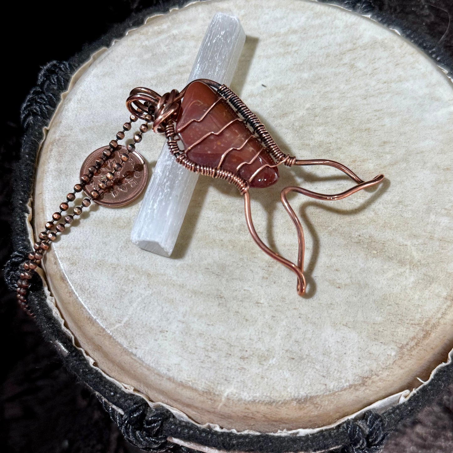 Handcrafted Red Jasper Mermaid Tail pendant resting on a selenite wand with a Canadian penny for scale, showcasing artisan reclaimed copper wirework and natural stone.