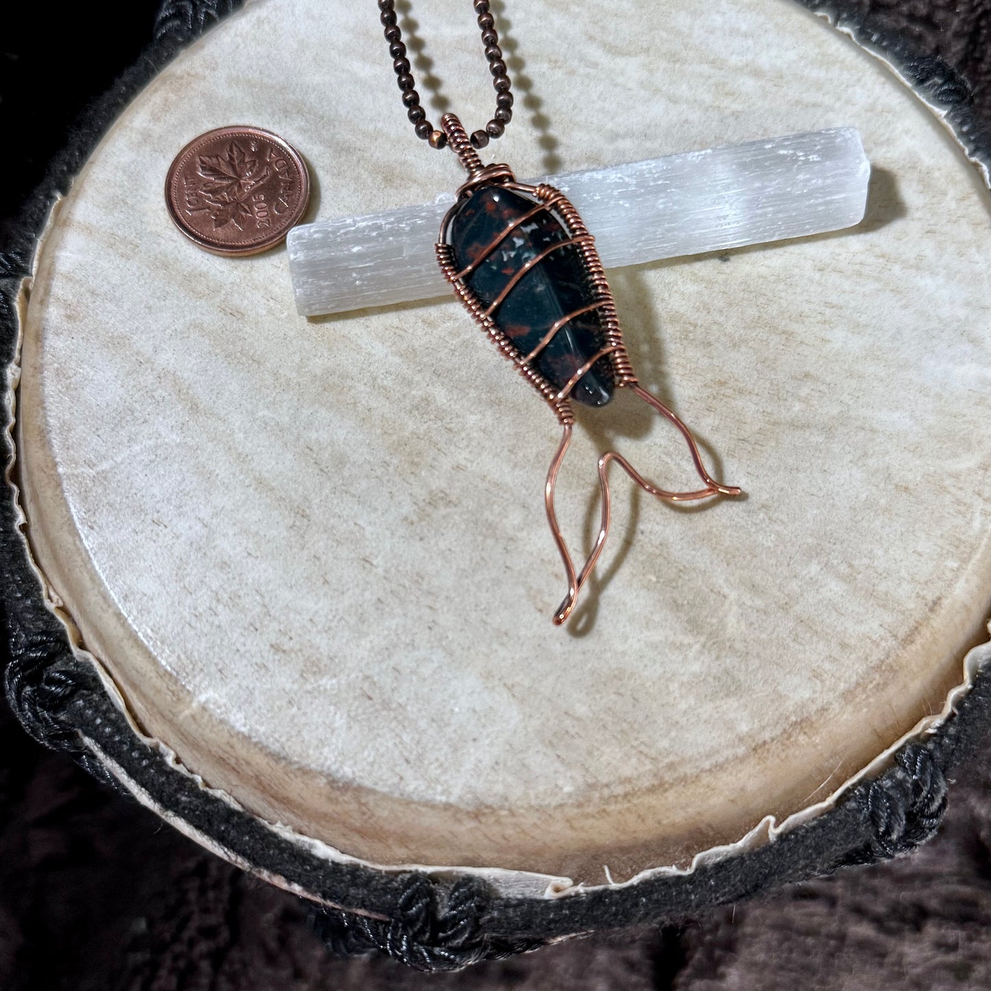 Bay of Fundy Bloodstone Mermaid Tail Necklace displayed with a Canadian penny for scale, highlighting reclaimed copper wirework and the stone's distinctive green and red colouring.