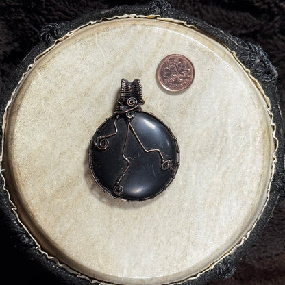 Reverse view of a round shungite pendant showing decorative copper wirework and a detailed patinated fine-wire frame.
