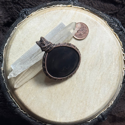Front display view of a dark shungite pendant wrapped in patinated copper with intricate fine-wire weaving and subtle natural pyrite flecks.