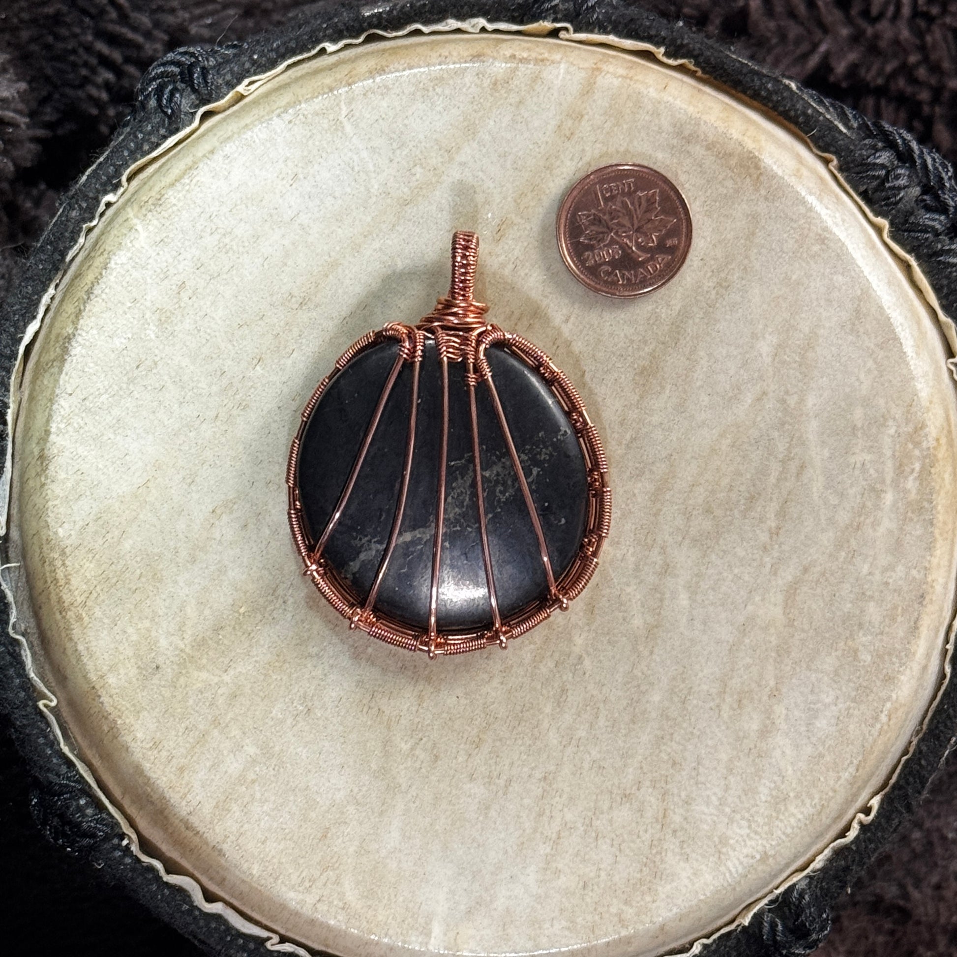 Back of a unisex shungite pendant showing radiating raw copper supports, detailed woven borders and the stone’s smooth rounded form.