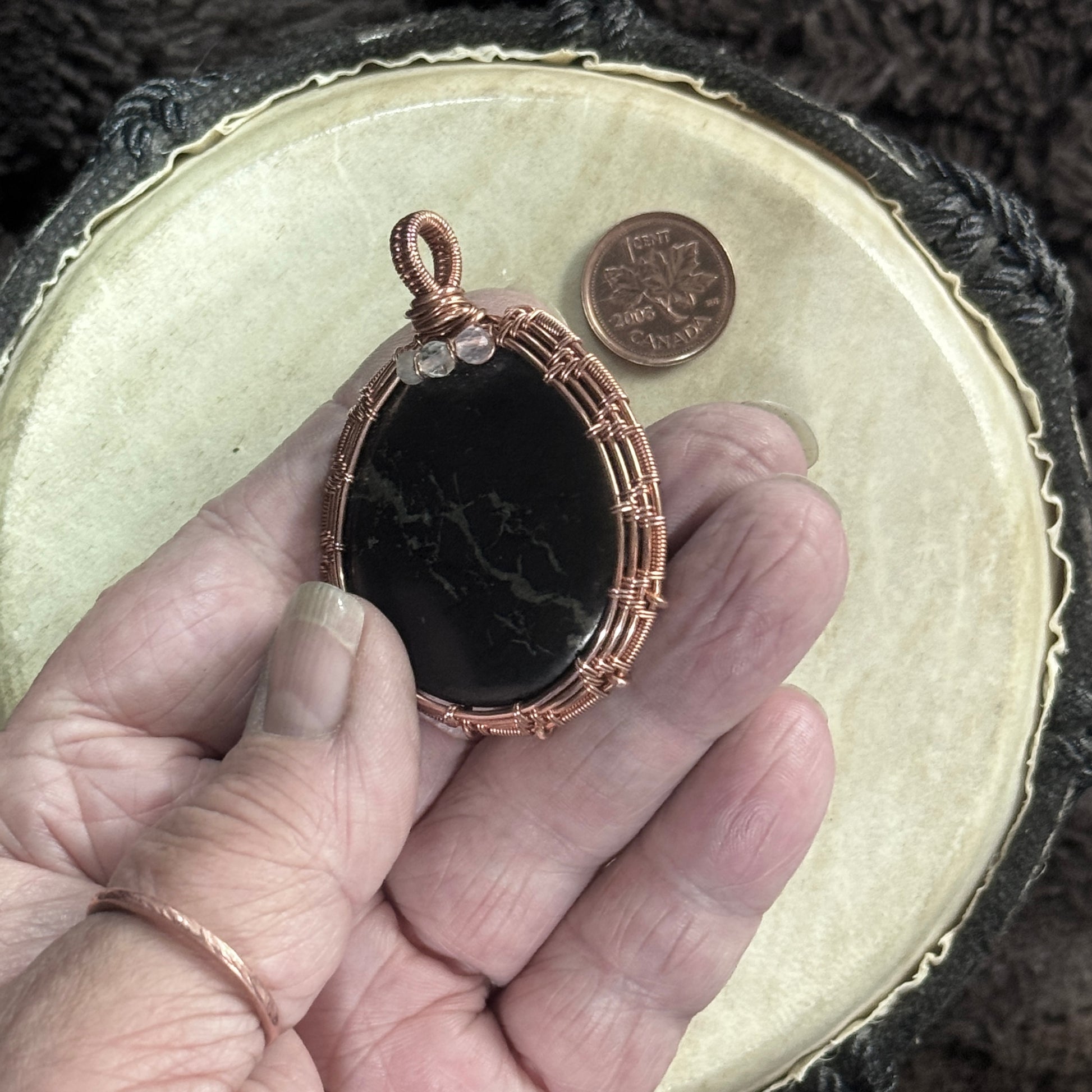 Hand-held front view of a tactile polished shungite pendant with a river-like pyrite seam, pale faceted fluorite and bright raw copper weaving.