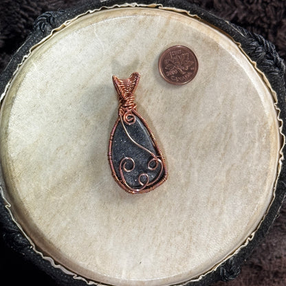 Side one of a reversible specular hematite pendant, sparkling charcoal-black within a handwoven raw copper frame with flowing spiral accents.